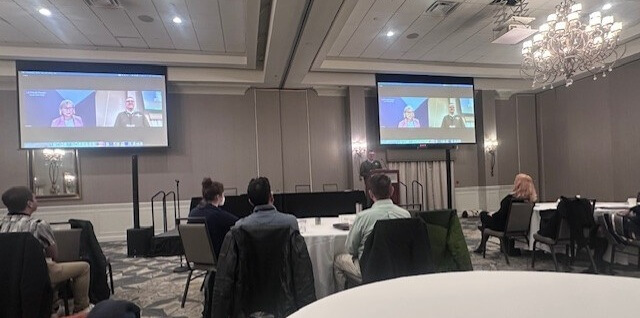 People gathered at round tables in a hotel conference room watching a virtual presenter on two monitors
