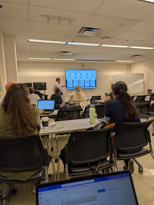 People sitting in a classroom while a person presents content on a screen at the front