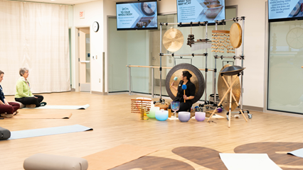 A woman seated on a mat with a gong and other instruments surrounding her as she leads other seated people in a sound immersion class