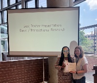 A picture of two women standing in front of a projector screen that reads Best Poster Presentation Basic / Translational Research. One woman holds a certificate.