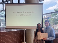 A picture of a woman next to a man holding a certificate. They are standing in front of a projector screen that reads Best Poster Presentation Clinical Research.