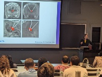 A picture of a woman standing at a podium giving a presentation next to a screen showing CT head images.